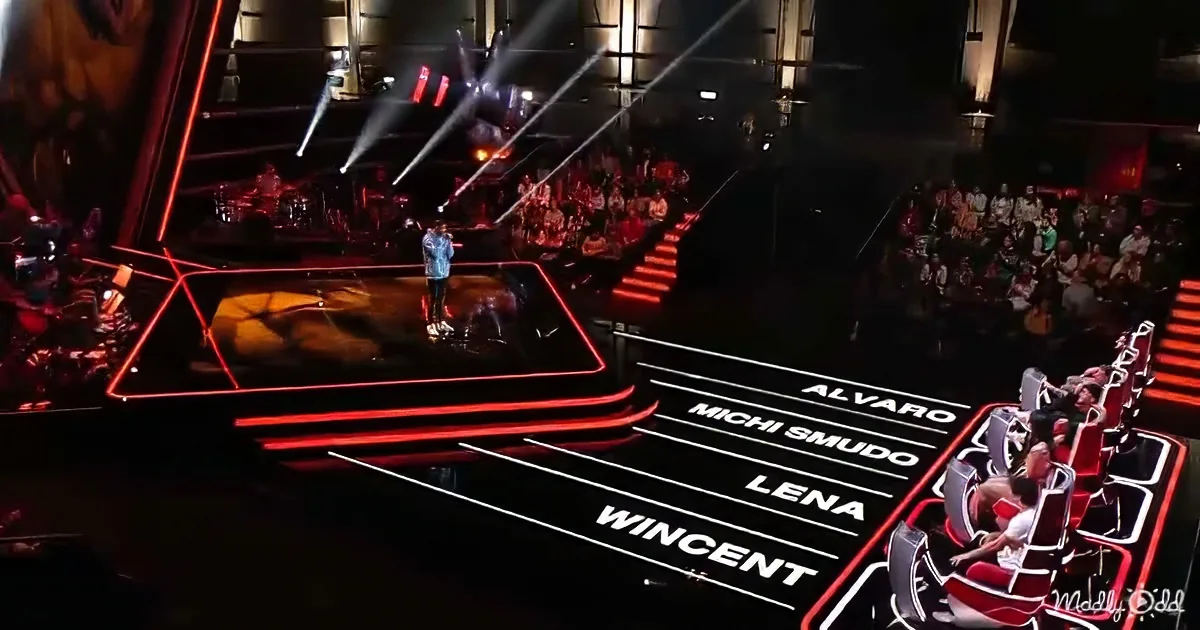 A contestant stands on a stage with red lighting, surrounded by judges' chairs and an audience.