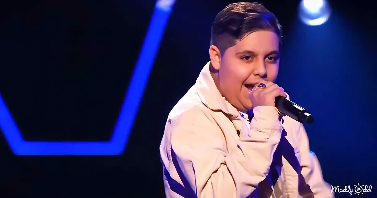 A person in a white jacket sings into a microphone on stage with blue lighting.
