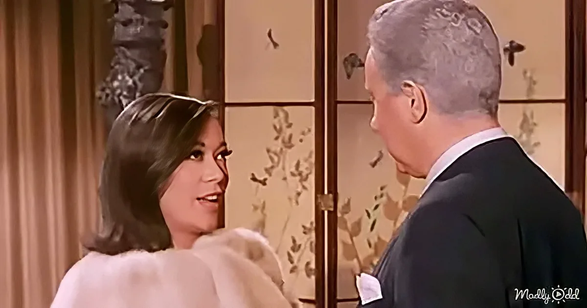 A woman in a white fur coat smiles at a man in a suit, both in a warmly lit room with floral wallpaper.
