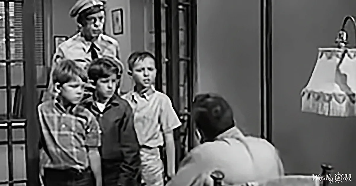 A man in a uniform stands above three boys, who appear to be in a room with a lamp and a window.