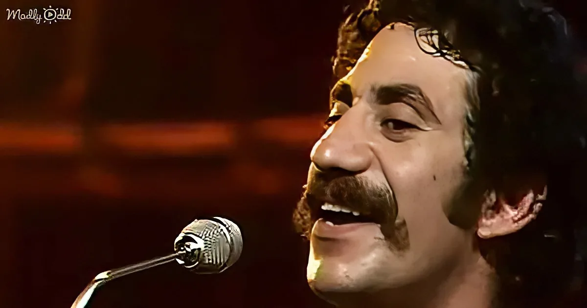 A man with curly hair and a mustache sings into a microphone, smiling with his eyes closed.