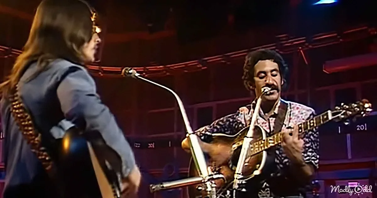 A musician in a patterned shirt plays a guitar on stage, with another person in a suit nearby.