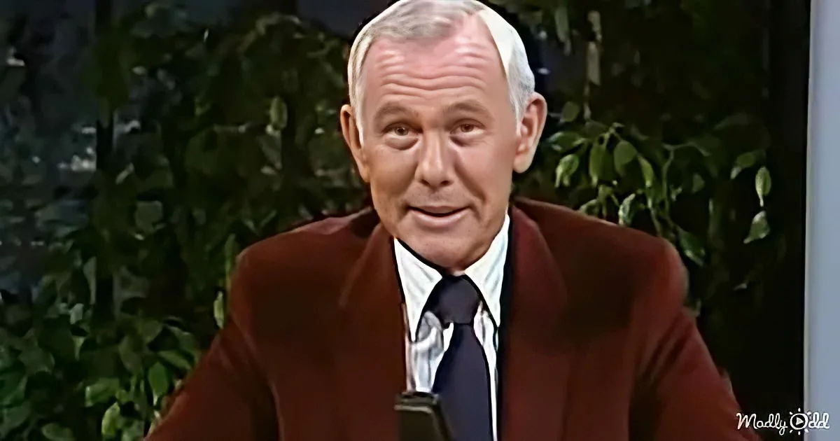 A man in a brown suit and white shirt sits in front of greenery, appearing to speak or present.