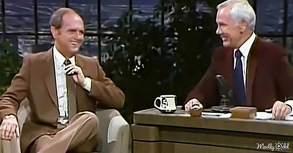 Two men in suits share a laugh on a talk show set with a cityscape backdrop.