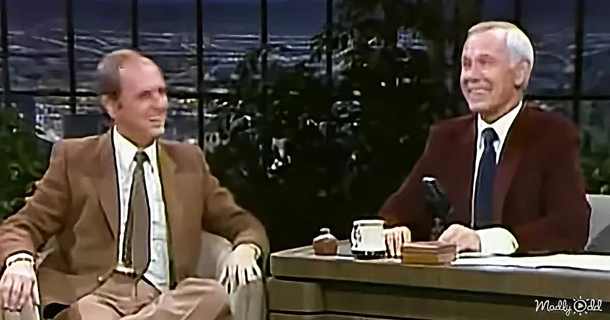Two men in suits sit on a talk show set, one smiling, against a nighttime cityscape backdrop.