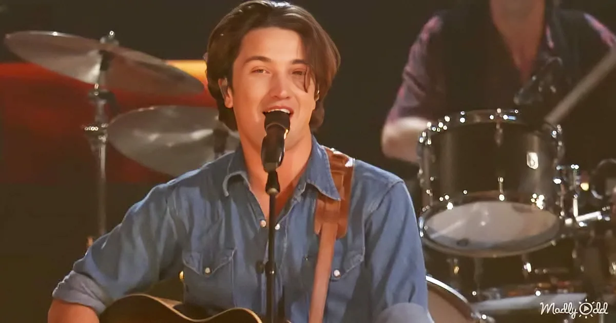 A man in a denim shirt sings into a microphone while playing guitar, with a drum set in the background.