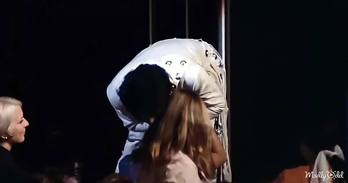 A person in a white, hooded outfit with a mask is being held by another person in a dark setting.
