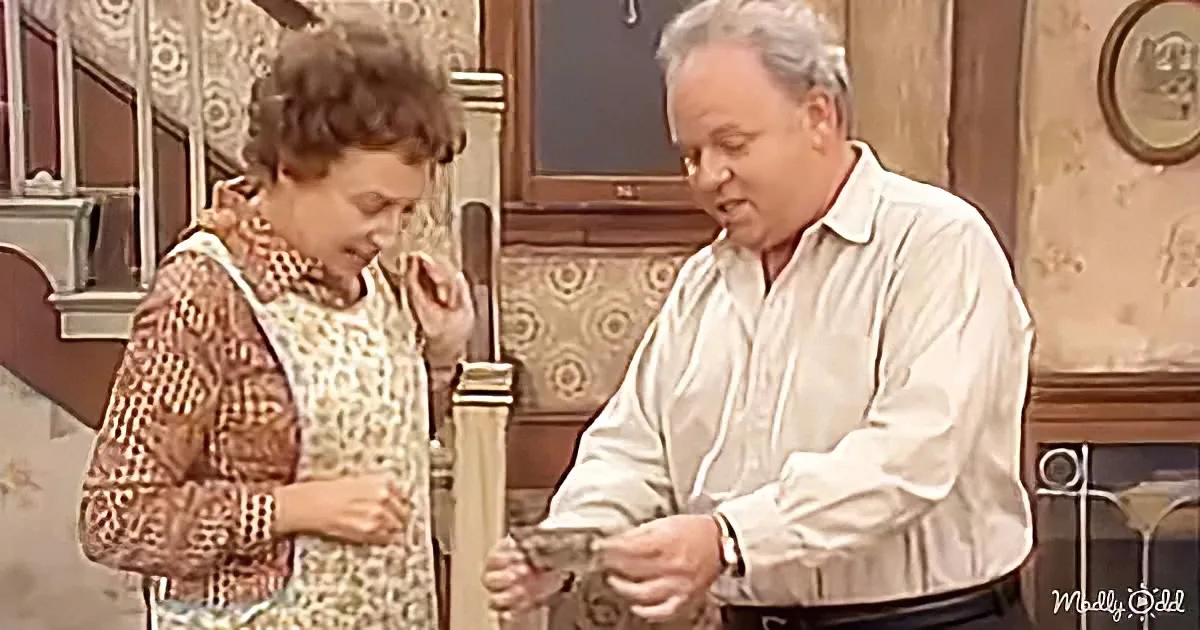 A man in a white shirt holds a small object, while a woman in a patterned dress looks down, both in a room with floral...
