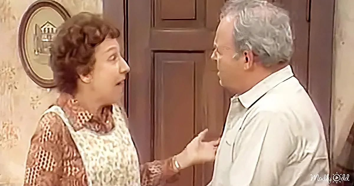 A woman in a patterned dress gestures while talking to a man in a white shirt in a room with a door and wall decoration.