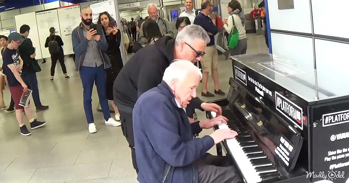 An older man in a blue jacket plays a piano in a busy public space, surrounded by people.