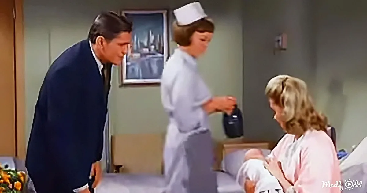 A man in a suit leans over a hospital bed where a woman holds a baby, with a nurse in the background.