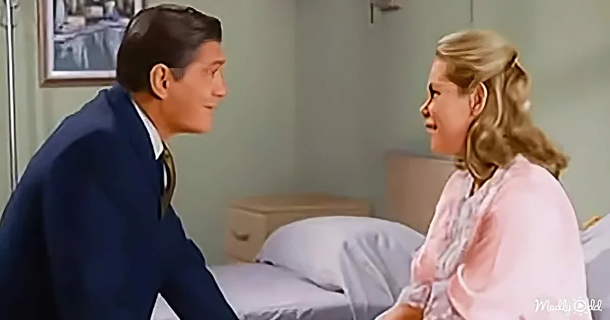 A man in a suit and a woman in a pink dress are sitting on a bed, facing each other, in a room with a painting on the wall.