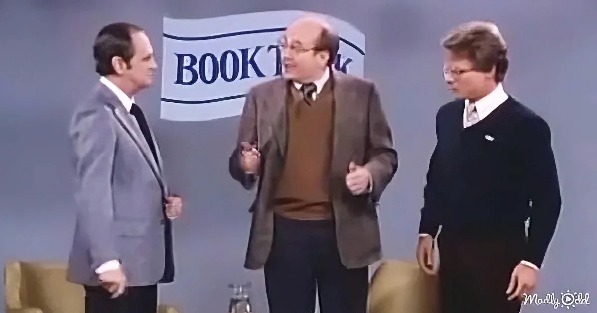 A man in a brown suit gestures animatedly while standing between two other men on a stage.