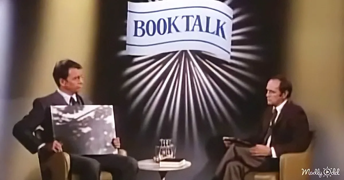 Two men in suits sit on stage with a "BOOK TALK" backdrop, one holding a book.
