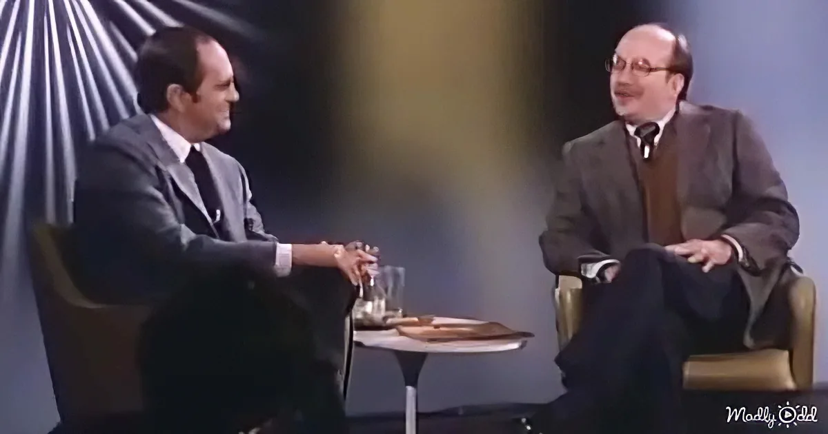 Two men in suits sit across from each other, smiling, with a table between them holding glasses and a plate.
