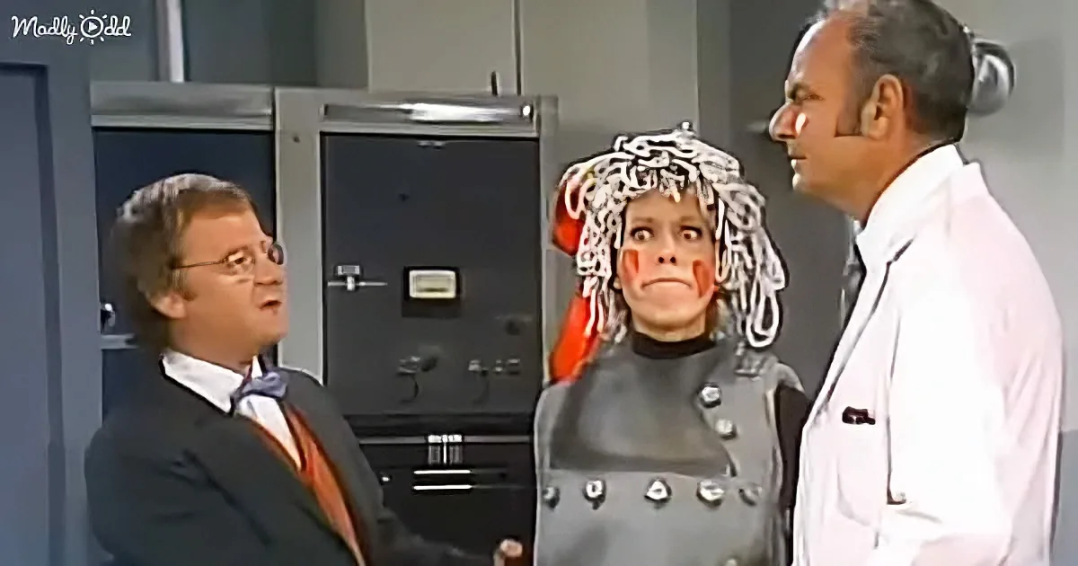 A person in a lab coat and another in a suit observe a third person with a strange head covering and red face paint.