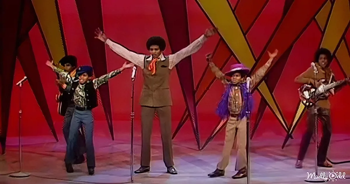 A man in a brown outfit with arms raised, surrounded by other performers in colorful costumes on a stage with a red and...