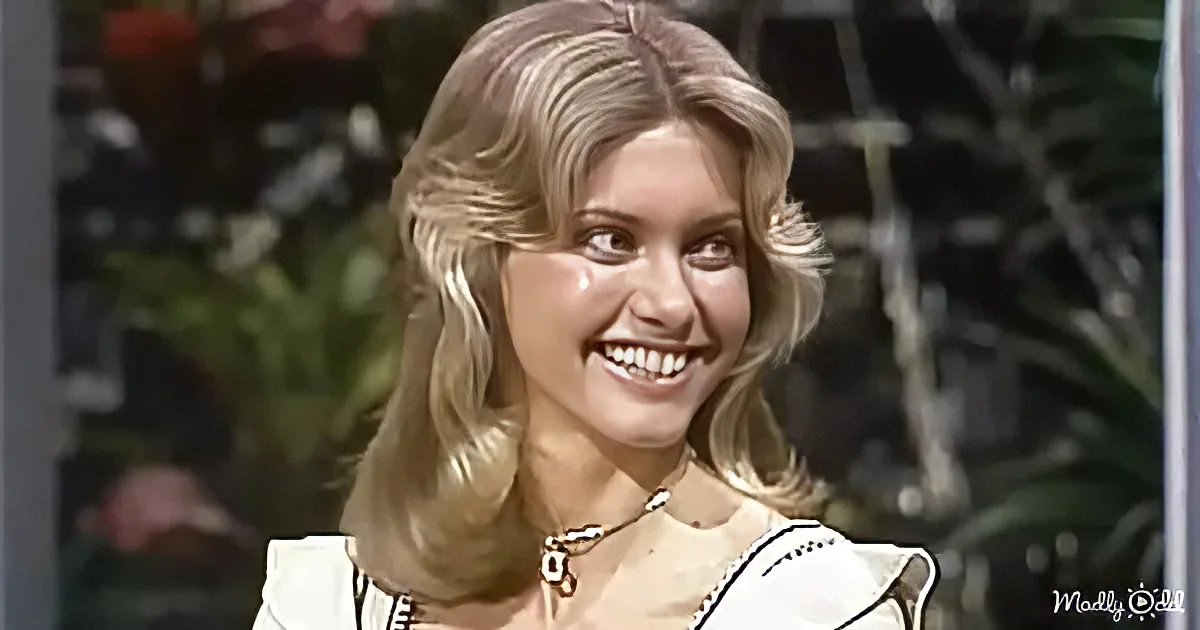 A smiling woman with blonde hair, wearing a white top and a necklace, stands in front of a blurred background with greenery.
