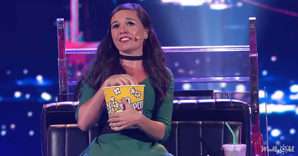 A woman in a green dress sits on a chair, holding a popcorn bucket, smiling, with a microphone on her ear.