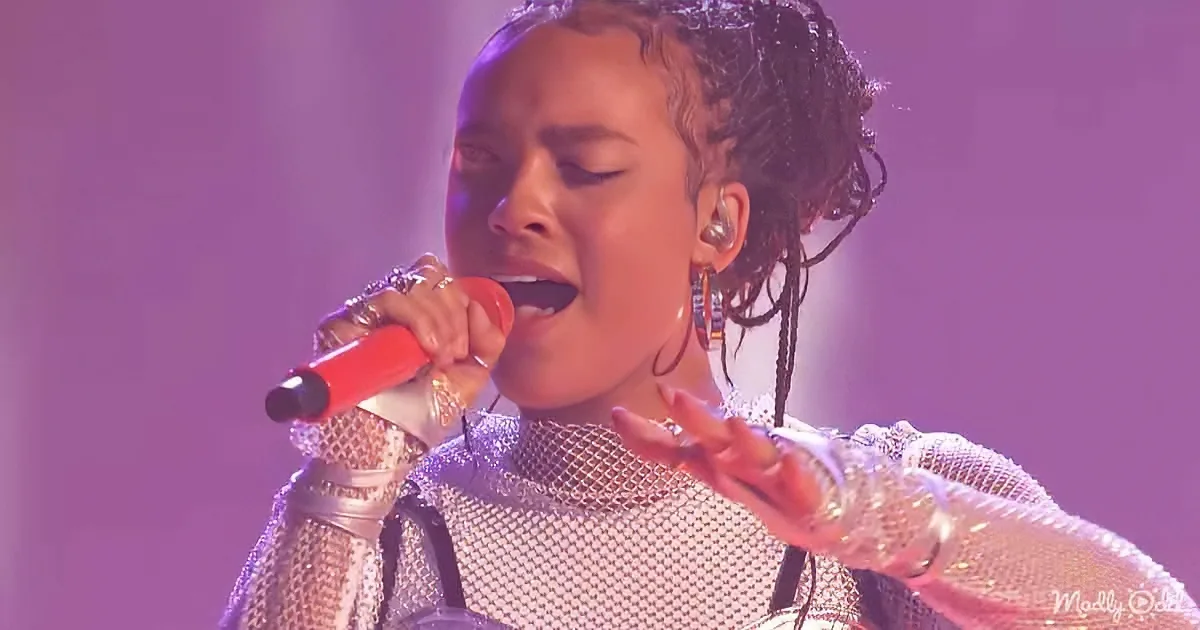 A person in a metallic outfit sings passionately into a red microphone, with a purple background.