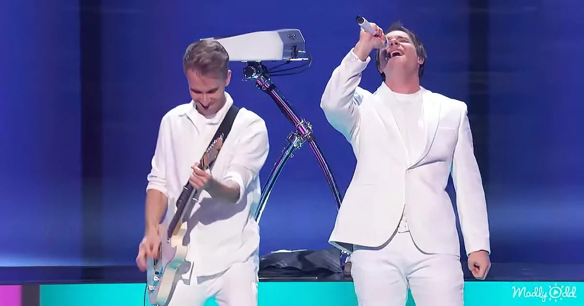 A man in white sings passionately while another plays guitar on stage.