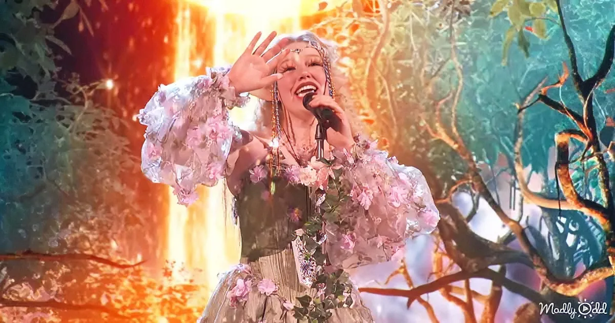 A performer in a floral dress sings passionately on stage with a vibrant, forest-themed backdrop.