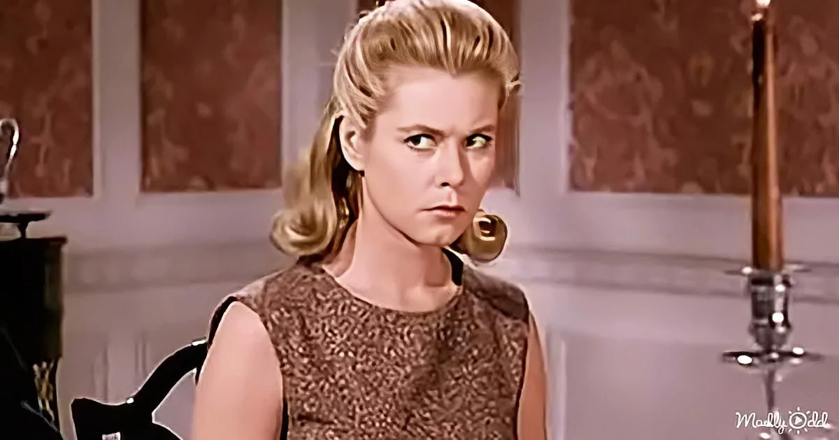 A woman with blonde hair in a 1960s-style dress sits in a room with patterned wallpaper and a candlestick.