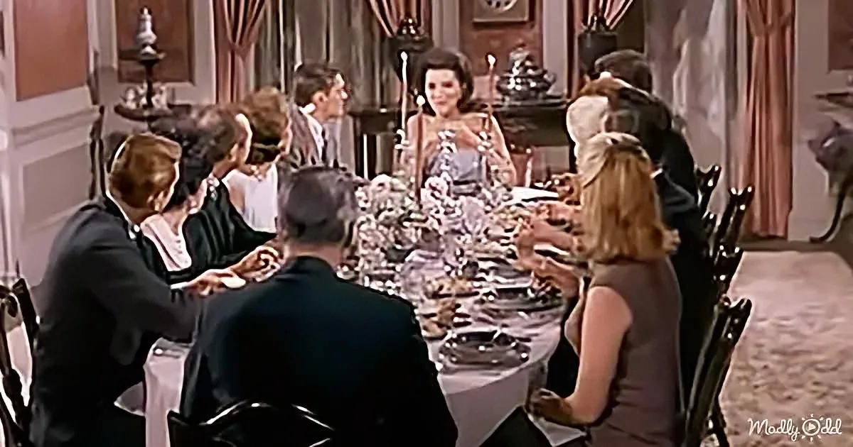 A woman in a formal dress stands at a dining table surrounded by guests in a sophisticated setting.