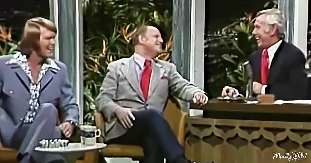 A man in a suit and red tie sits on a chair, laughing with others in a casual setting.