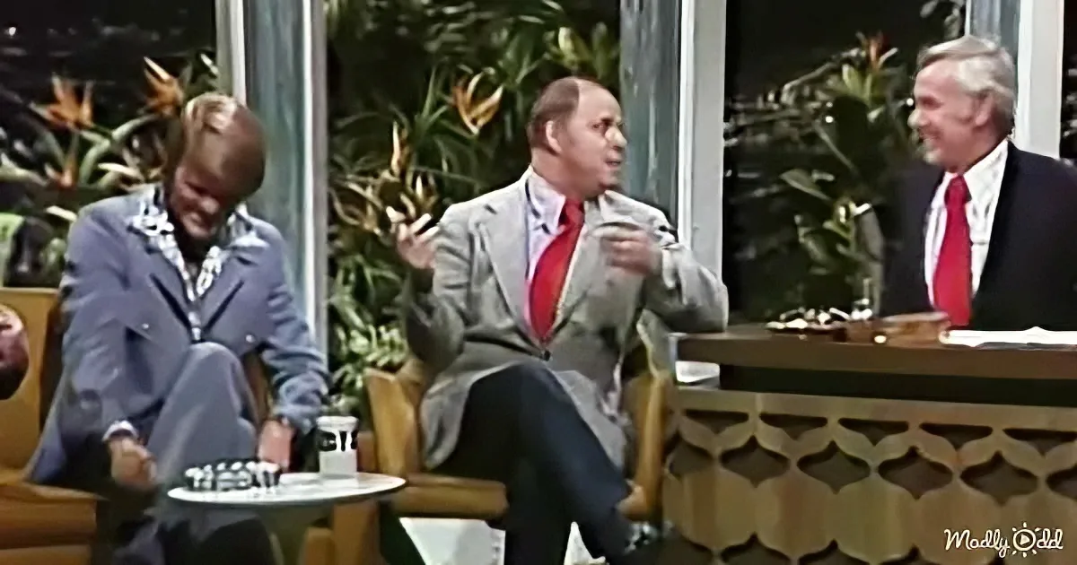 Three men in suits sit at a table, one laughing, another gesturing, in a setting with plants and a decorative wall.