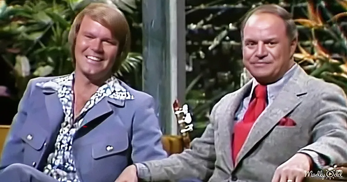 Two men in suits, one in a patterned shirt, sitting on a set with tropical plants in the background.