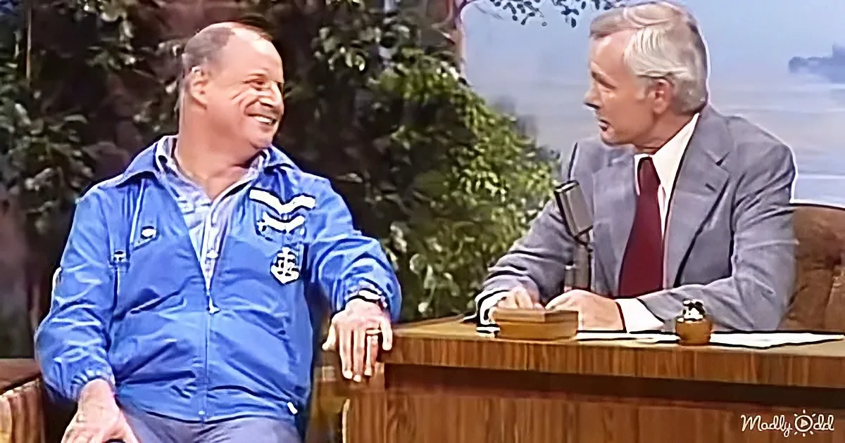 A man in a blue jacket leans on a desk, smiling, while another man in a suit sits behind it, holding a microphone.