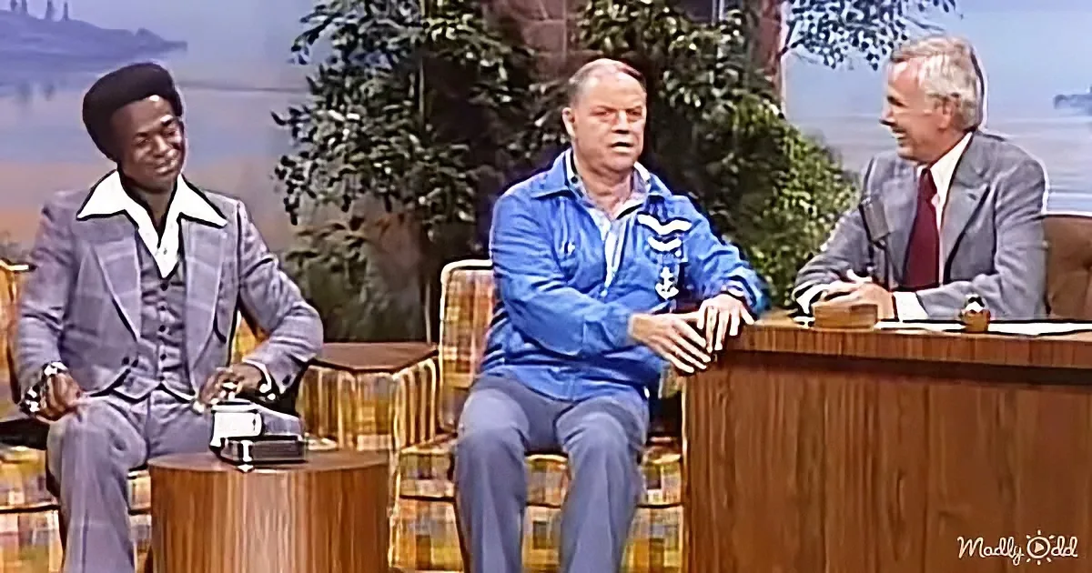 A man in a blue jacket sits on a chair, holding a small object, while another man in a suit and a third in a suit and tie...