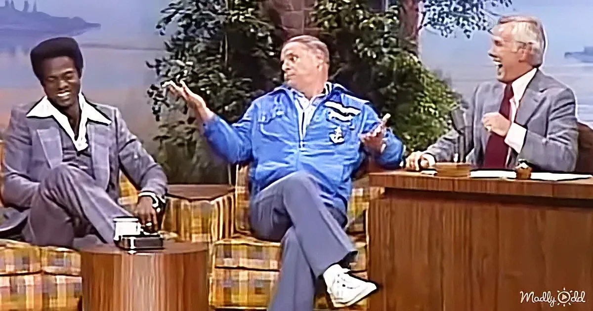 A man in a blue tracksuit gestures animatedly on a talk show set with two other men.