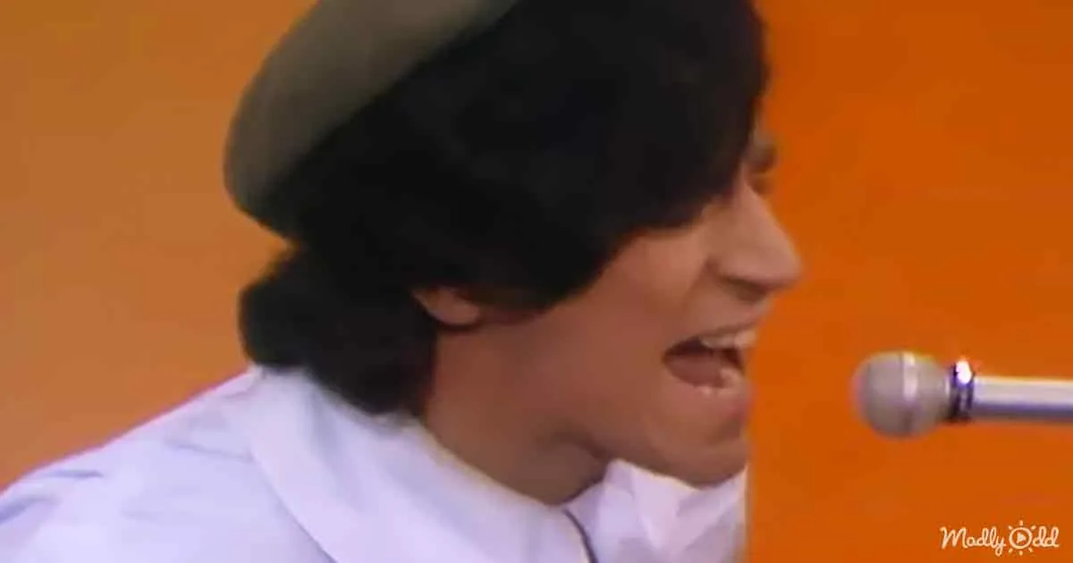 A person wearing a white robe and a black hat is speaking into a microphone.