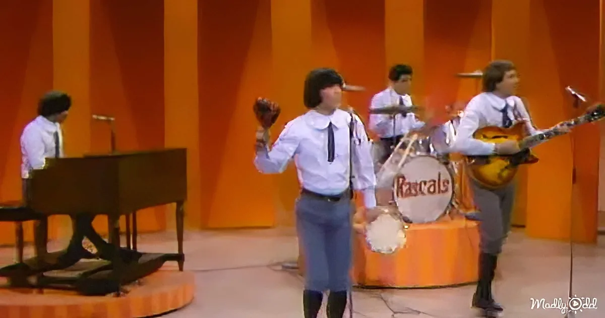 A person in a white shirt and tie plays a tambourine on stage with a band.