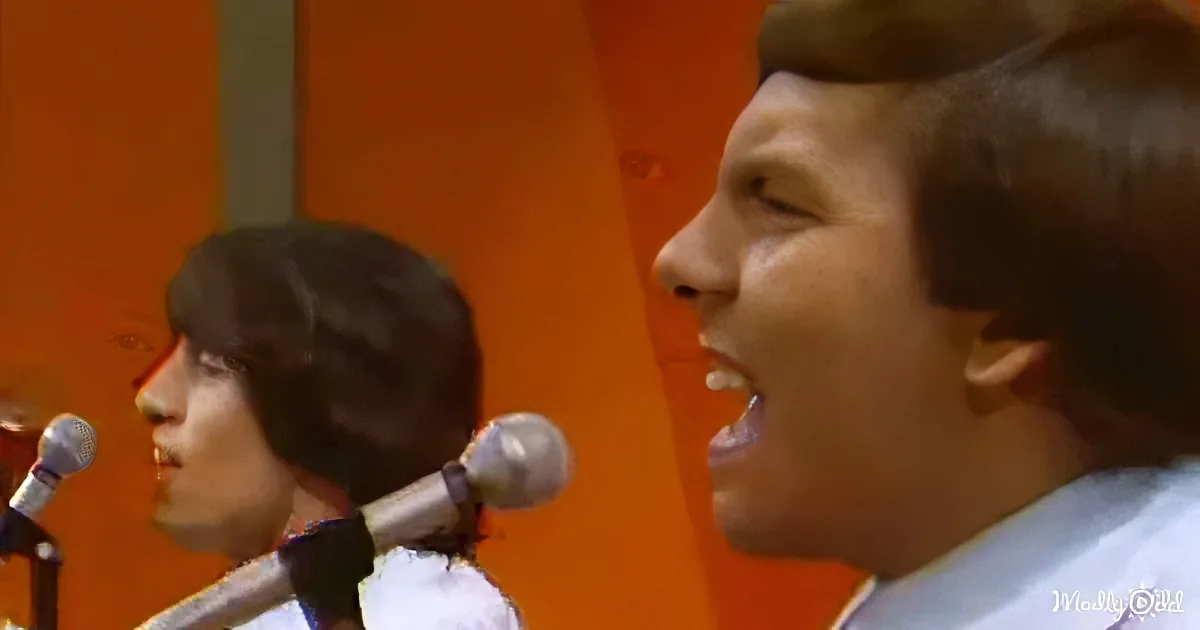 A young man with short hair sings into a microphone, with another person in the background.