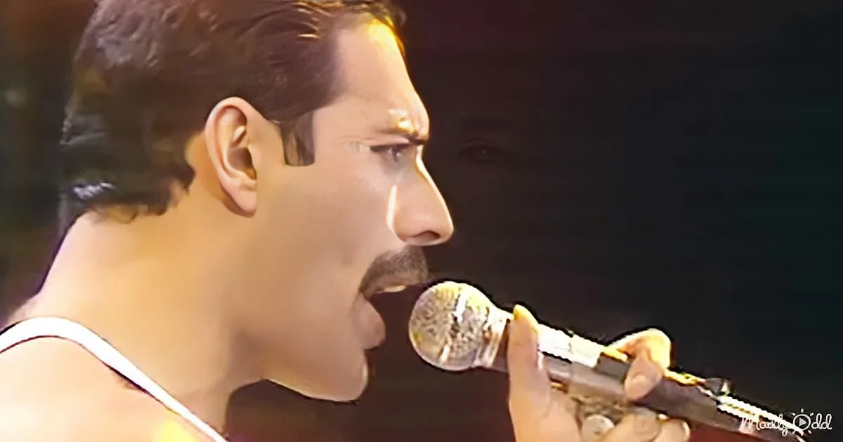 A man with short hair and a white tank top sings into a microphone, holding a guitar.