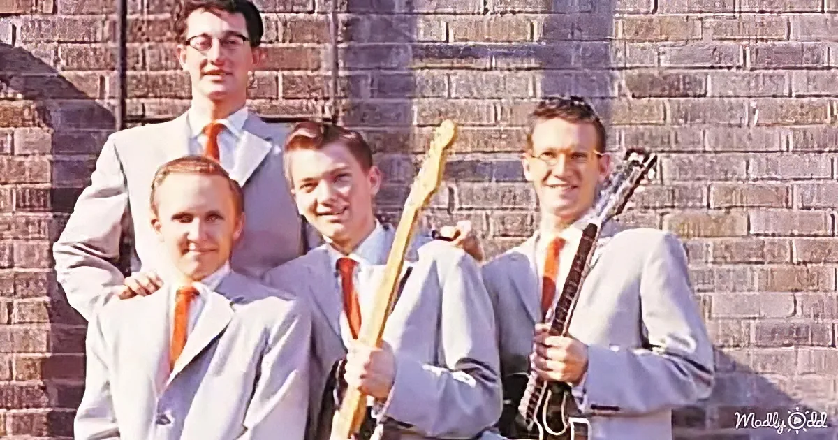 Four men in suits with guitars pose in front of a brick wall.