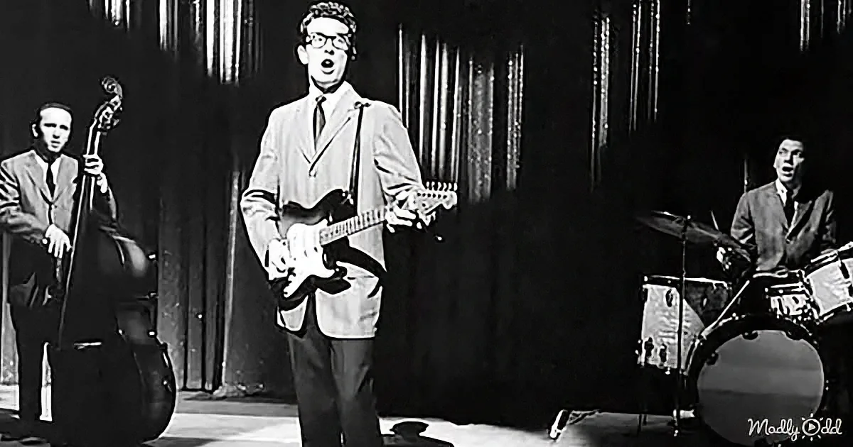 A man in a suit plays guitar and sings on stage with a bassist and drummer.