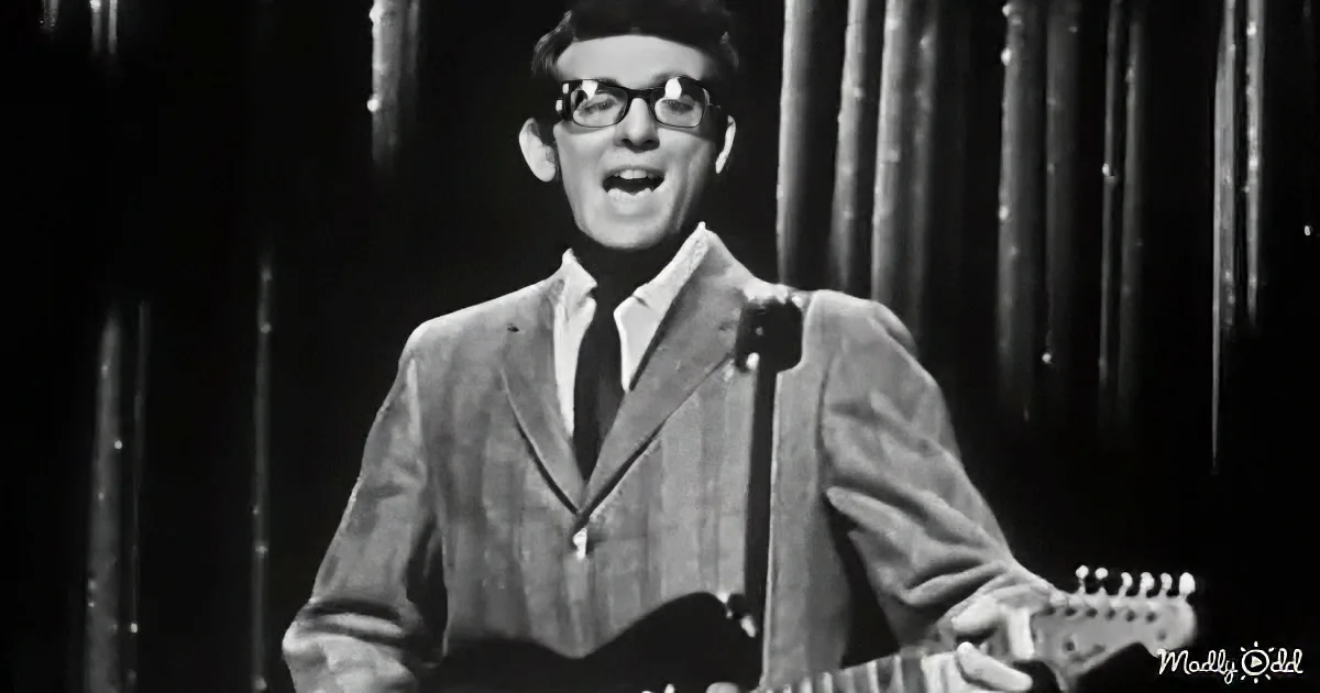 A man in a suit and tie sings into a microphone while holding a guitar.