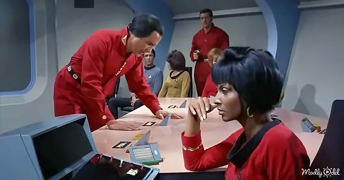 A person in a red uniform leans over a table with another person in a red uniform, surrounded by others in a futuristic...