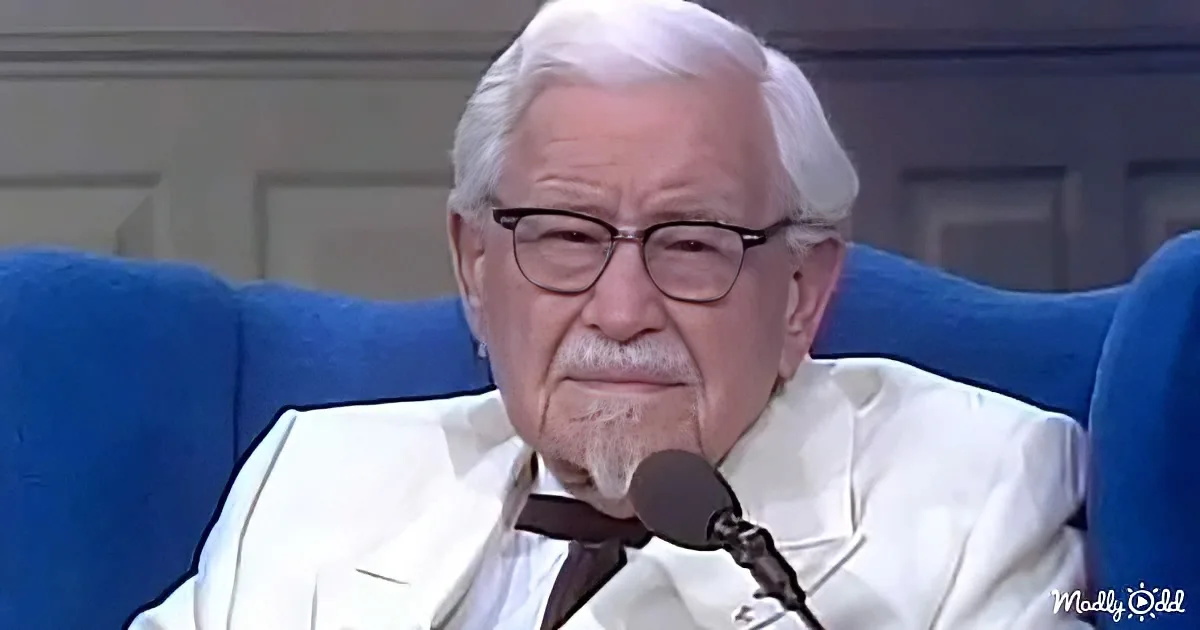 An elderly man with white hair and glasses, wearing a white suit, sits in a blue chair, speaking into a microphone.