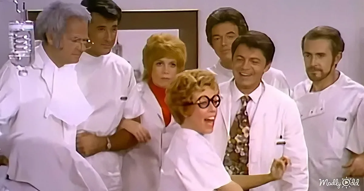 A woman in a lab coat and glasses is surrounded by a group of people in white lab coats in a laboratory setting.
