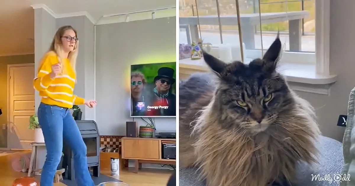 A woman dances in a living room with a cat sitting on a couch in the background.