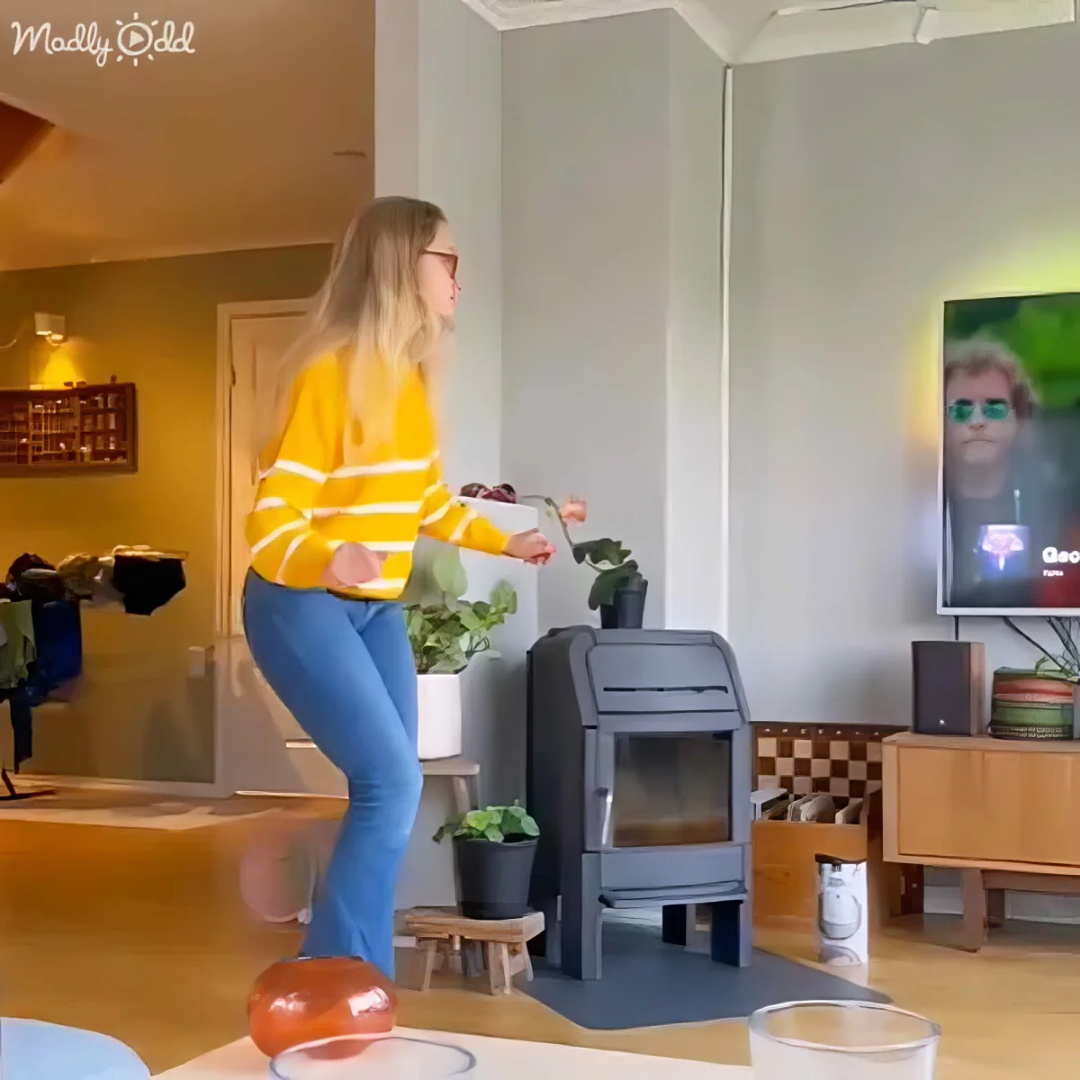 A woman in a yellow sweater balances on a glass ball in a living room with a TV showing a person.