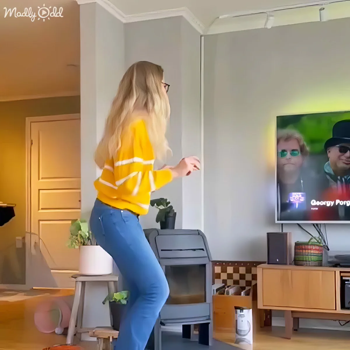 A woman in a yellow sweater dances in front of a TV showing a man in sunglasses.