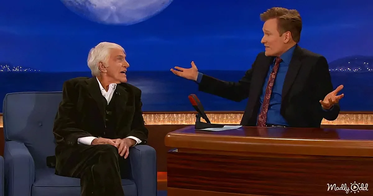 Conan O'Brien gestures while interviewing a guest on a late-night talk show set.