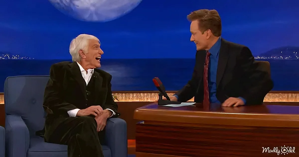 Two men are seated on a talk show set, one in a suit laughing, the other in a dark suit with a red tie.
