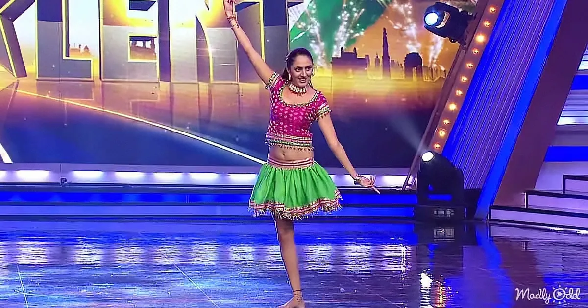 A woman in a colorful traditional outfit dances on a stage with bright lights and a cityscape backdrop.